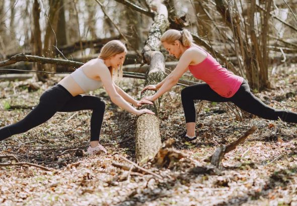 Side view of young female athletes in activewear stretching legs together using fallen tree during fitness workout in spring forest adhering to healthy lifestyle